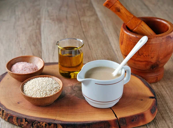Tahini omáčka v džbánu se ingrediencemi. Sezamová semena, olivový olej a sůl na dřevěné desce. — Stock fotografie
