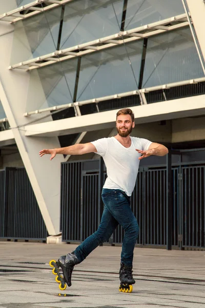 Hombre barbudo en patines de pie en el fondo del edificio. Joven hombre en forma con camisetas blancas y pantalones vaqueros en patines de ruedas cabalgando al aire libre después de la lluvia . — Foto de Stock