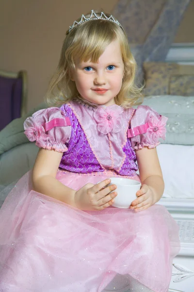 Pequena menina princesa feliz em vestido rosa e coroa em seu quarto real sentado na cadeira e segurando xícara de bebida . — Fotografia de Stock