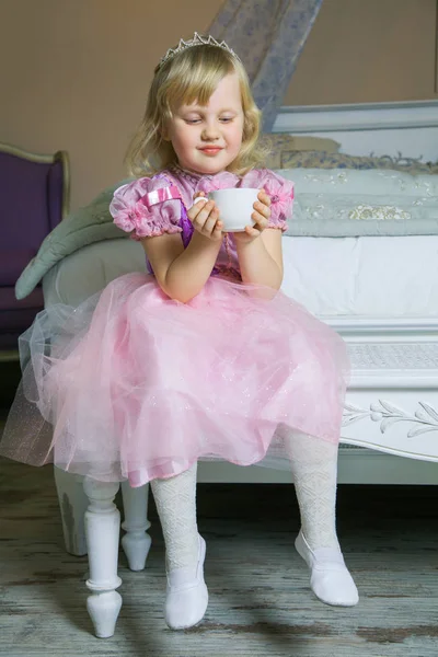 Šťastná princezna holčička v růžových šatech a koruny v její královské místnosti, sedí na židli a drží šálek nápoje. — Stock fotografie