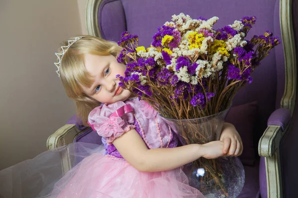 Šťastná princezna holčička v růžových šatech a koruny v její královské místnosti, sedí na židli a drží váza s květinami. — Stock fotografie