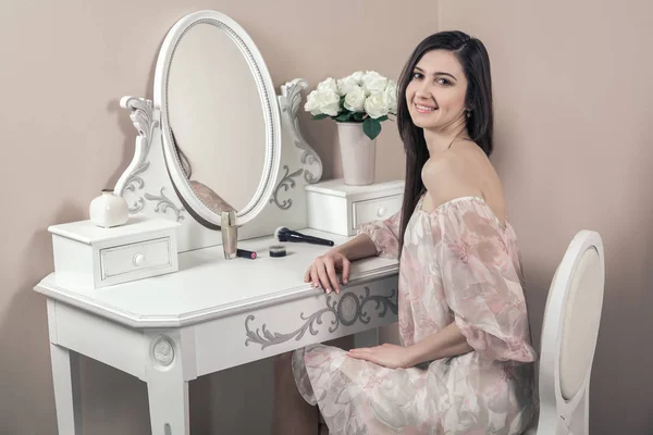 Vacker glad kvinna med rosa klänning och långa svarta hår i hennes rum nära hennes toalettbord poserar innan part. — Stockfoto