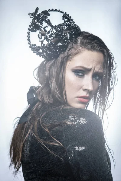 Mode-Schönheitsporträt einer jungen schönen jungen Frau mit Make-up und Sommersprossen im Gesicht mit schwarzer Krone auf dem Kopf und schwarzem Kleid. mit Licht von hinten auf hellem Hintergrund. — Stockfoto