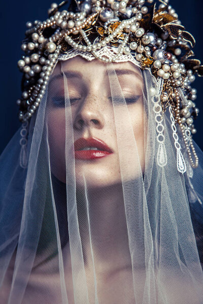 fashion beauty portrait of young beautiful young woman with makeup and freckles on her face and pearl headpiece on her head and white tulle in front of her face on dark blue background. 