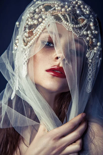 Mode Schönheit Porträt der jungen schönen jungen Frau mit Make-up und Sommersprossen im Gesicht und Perlenkopf auf dem Kopf und weißem Tüll vor ihrem Gesicht auf dunkelblauem Hintergrund. — Stockfoto