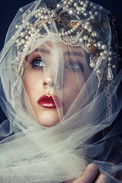 Retrato de belleza de moda de una joven hermosa joven con maquillaje y pecas en su cara y casco de perlas en su cabeza y tul blanco en frente de su cara sobre fondo azul oscuro . — Foto de Stock