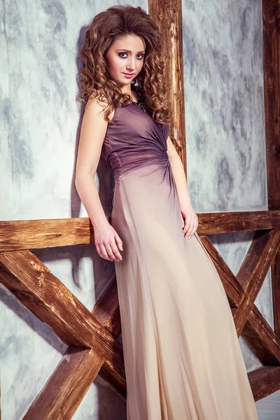 Mode-Model mit langem Kleid und lockiger Frisur und Make-up posiert in der Nähe der Wand mit Holzstange. Studioaufnahme. — Stockfoto