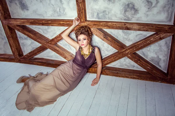 Модна модель з довгим платтям і кучерявою зачіскою і макіяжем позує біля стіни з дерев'яним полюсом. студійний знімок . — стокове фото