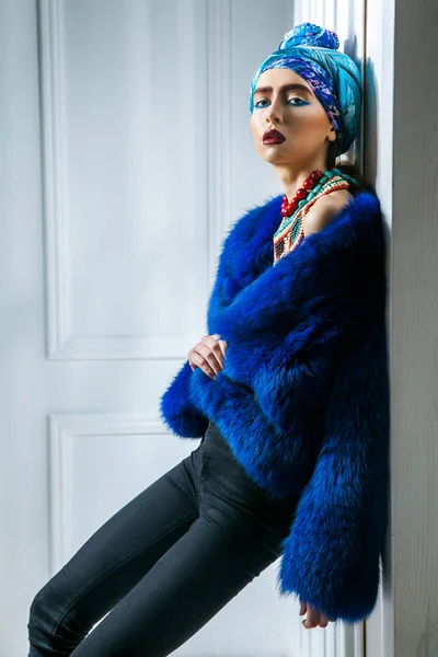 Schönheitsporträt des Modells mit farbiger Kopfbedeckung, blauem Pelzmantel rote Augenbrauen und Lippen Make-up und Halskette. Studioaufnahme in der Nähe von Fenstern und weißer Wand — Stockfoto