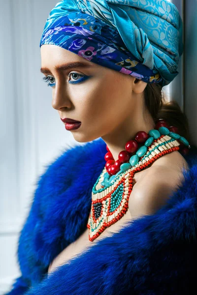 Красивий портрет моделі моди з кольоровим головним убором, синьою шубою червоні брови і макіяж губ і намисто. студійний знімок біля вікон і білої стіни — стокове фото
