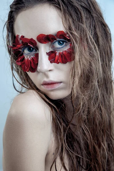 Retrato de belleza de la joven modelo de moda con pelos húmedos y pétalos de flores rojas alrededor de sus ojos. estudio de tiro sobre fondo azul —  Fotos de Stock