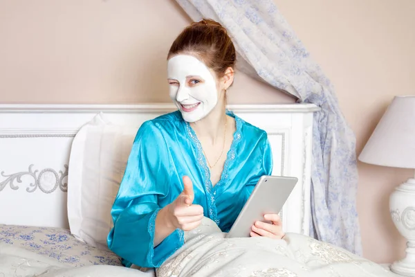 Bella donna con camicia da notte verde e maschera facciale bianca poggiata sul letto e che tiene tablet prima di dormire — Foto Stock