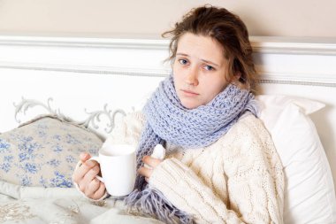 Çay ile hasta bir kadın. Bir fincan çay odasında yatakta otururken tutan örme eşarp mavi genç sinirli kadın portre görüntüsü. kameraya üzüntü ile seyir.