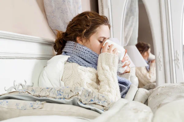 Donna malata con una tazza di tè. Immagine da primo piano di una giovane donna frustrata in una sciarpa blu lavorata a maglia che tiene e beve una tazza di tè mentre è seduta nel letto della sua stanza . — Foto Stock