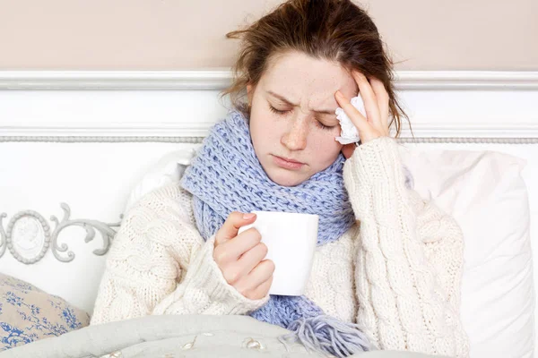 Donna malata con una tazza di tè. Immagine da primo piano di una giovane donna frustrata in sciarpa blu lavorata a maglia che tiene una tazza di tè mentre è seduta nel letto della sua stanza . — Foto Stock