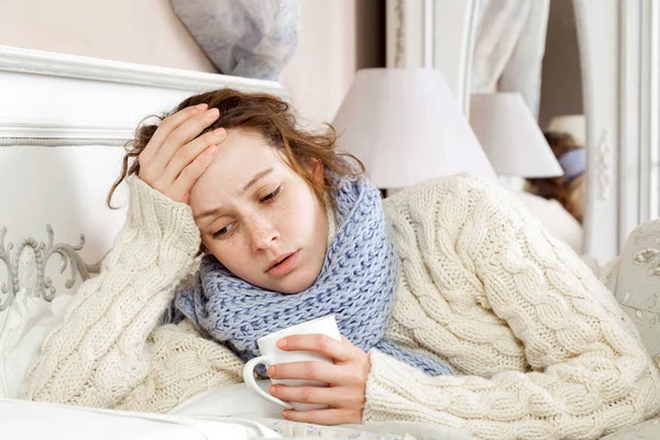 Sick woman with cup of tea. Closeup image of young frustrated sick woman in knitted blue scarf holding a cup of tea while lying in bed. hand on head, sad and feeling bad. — Stock Photo, Image