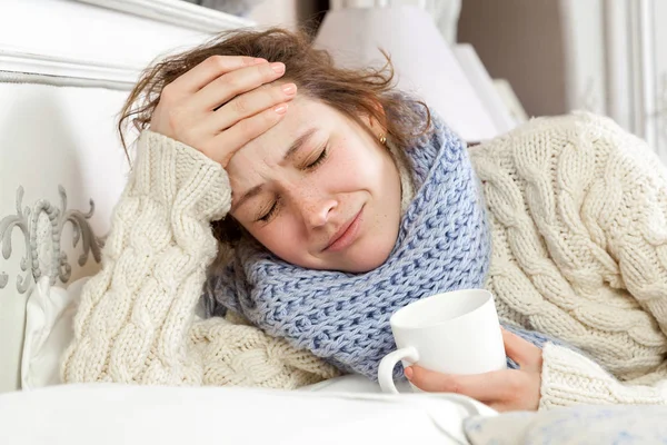 Wanita sakit dengan secangkir teh. Gambar close-up dari wanita muda yang sakit frustrasi dalam syal biru rajutan memegang secangkir teh sambil berbaring di tempat tidur. tangan di atas kepala, sedih dan menutup mata . — Stok Foto