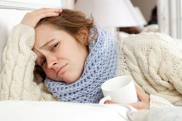 Sick woman with cup of tea. Closeup image of young frustrated sick woman in knitted blue scarf holding a cup of tea while lying in bed. hand on head, sad and looking at camera. — Stock Photo, Image