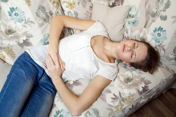 Ung smuk pige med mavesmerter liggende på sofaen - Stock-foto