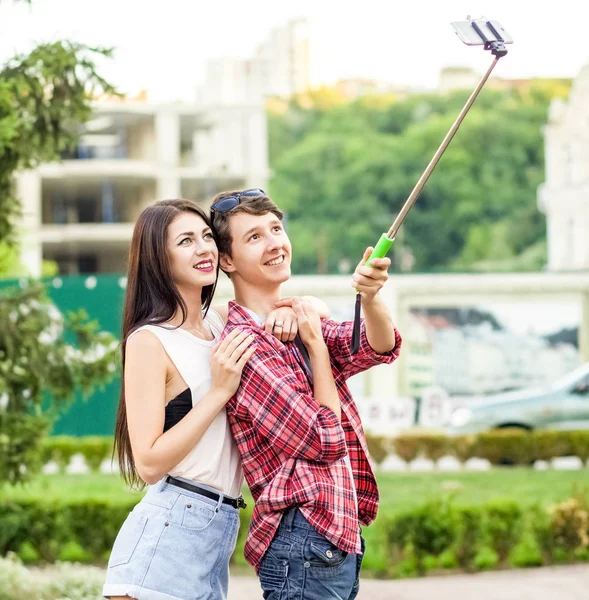 Feliz joven pareja de turistas tomando una selfie con teléfono inteligente en el monópodo en la ciudad. El hombre está sosteniendo el palo y disparando mirando el teléfono con felicidad —  Fotos de Stock