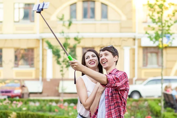 Feliz joven pareja de turistas tomando una selfie con teléfono inteligente en el monópodo en la ciudad. El hombre está sosteniendo el palo y disparando mirando el teléfono con felicidad —  Fotos de Stock