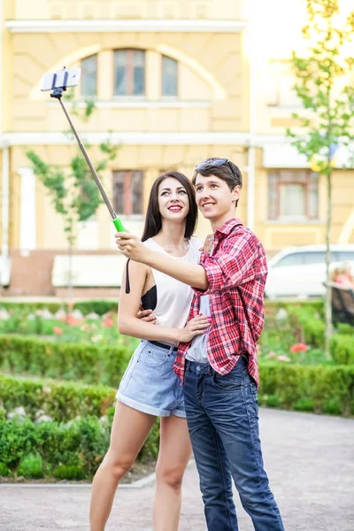 Feliz joven pareja de turistas tomando una selfie con teléfono inteligente en el monópodo en la ciudad. El hombre está sosteniendo el palo y disparando mirando el teléfono con felicidad —  Fotos de Stock