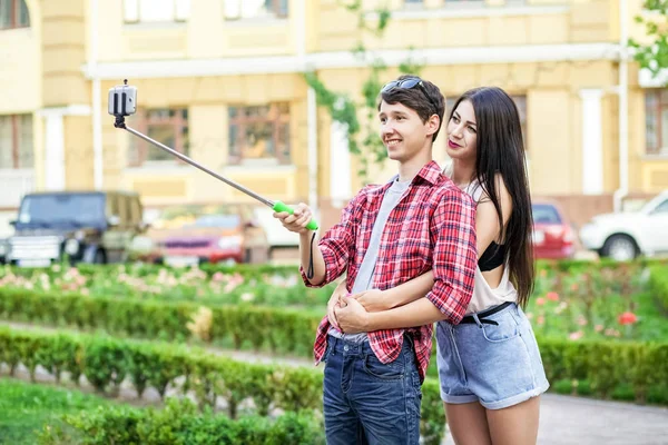 Feliz joven pareja de turistas tomando una selfie con teléfono inteligente en el monópodo en la ciudad. El hombre está sosteniendo el palo y disparando mirando el teléfono con felicidad — Foto de Stock