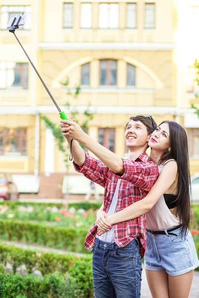 Feliz joven pareja de turistas tomando una selfie con teléfono inteligente en el monópodo en la ciudad. El hombre está sosteniendo el palo y disparando mirando el teléfono con felicidad —  Fotos de Stock