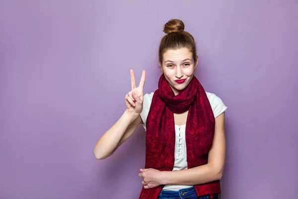 Porträtt av vackra unga glada leende kvinna, visar två fingrar eller seger gest, klädd vit t-shirt och röda halsduk på lila bakgrund. — Stockfoto