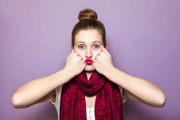 Retrato de una joven linda mujer con bufanda roja y pecas en su cara soplando beso y mirando a la cámara en el fondo púrpura —  Fotos de Stock