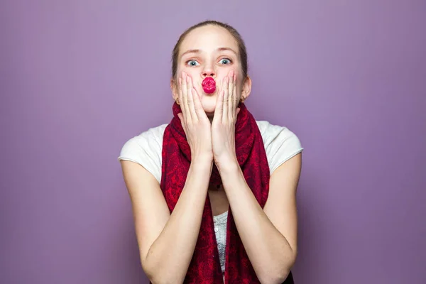 Retrato de una joven linda mujer con bufanda roja y pecas en su cara soplando beso y mirando a la cámara en el fondo púrpura — Foto de Stock