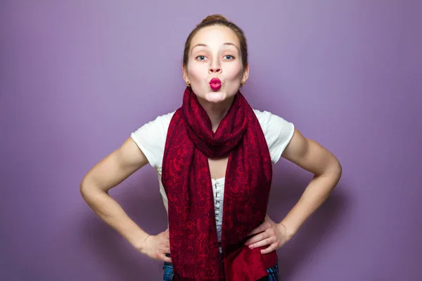 Porträt einer jungen hübschen Frau mit rotem Schal und Sommersprossen im Gesicht, die einen Kuss bläst und auf lila Hintergrund in die Kamera blickt — Stockfoto