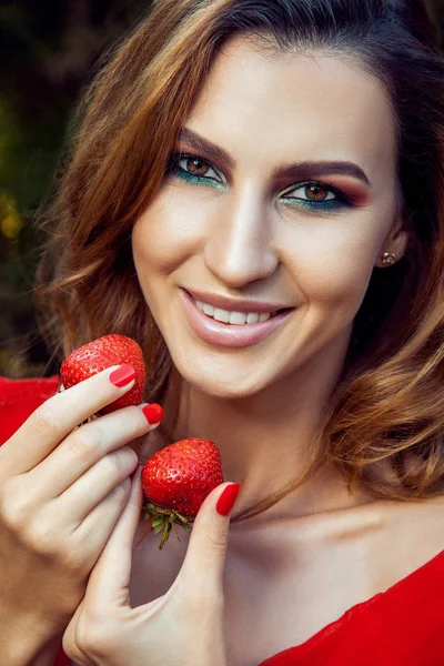 Menina engraçada feliz bonita nova com vestido vermelho e maquiagem segurando morango no verão no parque. estilo de vida saudável, dieta beleza e felicidade conceito . — Fotografia de Stock