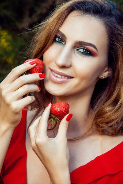 Krásná mladá šťastná legrační dívka s červenými šaty a make-up drží jahody v létě v parku. zdravý životní styl, strava krásy a štěstí koncept. — Stock fotografie