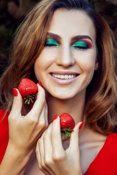Young beautiful happy funny girl with red dress and makeup holding strawberry in summertime in the park. healthy lifestyle, diet beauty and happiness concept. — Stock Photo, Image