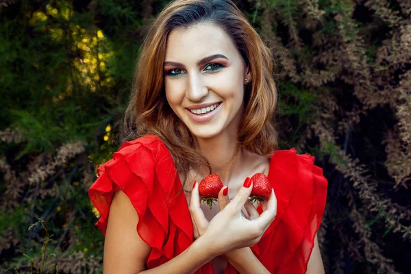 Menina engraçada feliz bonita nova com vestido vermelho e maquiagem segurando morango no verão no parque. estilo de vida saudável, dieta beleza e felicidade conceito . — Fotografia de Stock