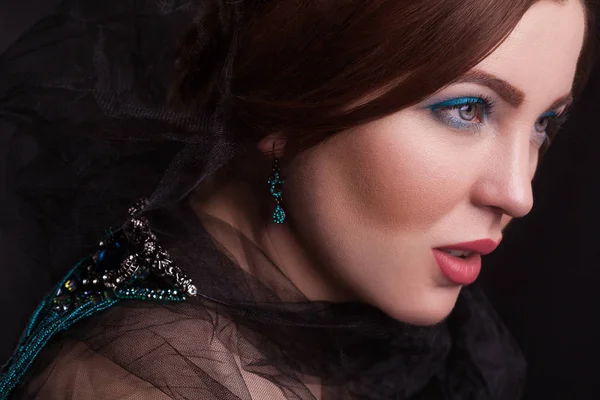 Portrait of beautiful brunette fashion model with black veil and blue jewelry and makeup and red lips in black bakground. — Stock Photo, Image