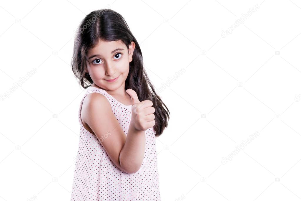 Beautiful happy little girl with long dark hair and dress looking at camera with thumbs up. studio shot, isolated on white background.