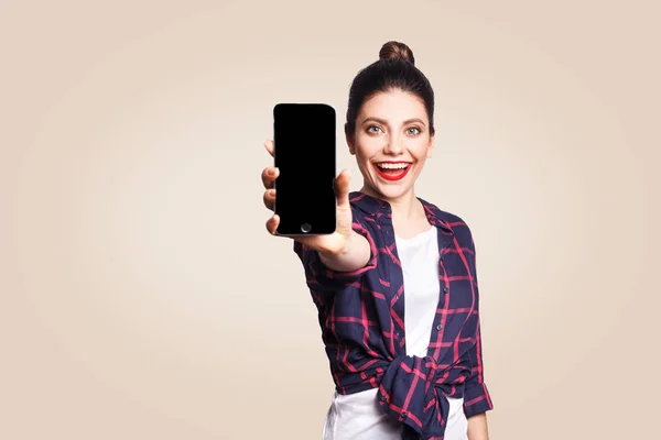Junge schöne Frau in lässigem Stil mit Handy, das in die Kamera schaut und das Handydisplay zeigt. Studioaufnahme auf beigem Hintergrund. — Stockfoto