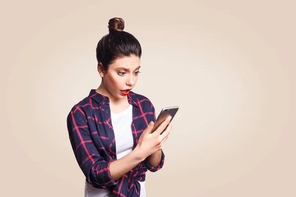 Mooie jongedame in casual stijl bedrijf telefoon en verrast. camera kijken met schok. studio opname op beige achtergrond. — Stockfoto