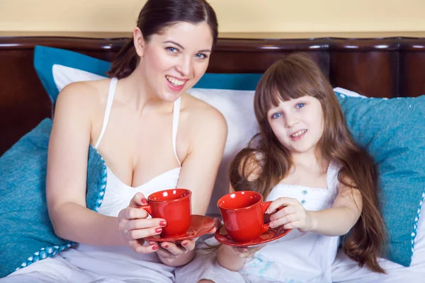 Feliz madre e hija divirtiéndose y sosteniendo la taza y descansando en la cama — Foto de Stock