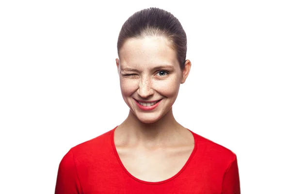 Retrato de una divertida mujer positiva guiñando el ojo en camiseta roja con pecas. mirando a la cámara con una sonrisa dentada, filmado en el estudio. aislado sobre fondo blanco . —  Fotos de Stock