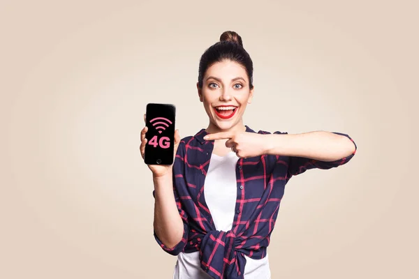 Mujer hermosa joven en estilo casual sosteniendo el teléfono mirando a la cámara y mostrando Internet 4G en la pantalla del teléfono con el dedo. estudio rodaje sobre fondo beige . — Foto de Stock