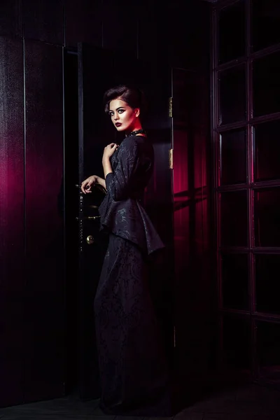 在经典的黑色礼服、 化妆和发型接近黑暗的门站和摆姿势和红光从她的窗口发光美丽时装模特的肖像. — 图库照片