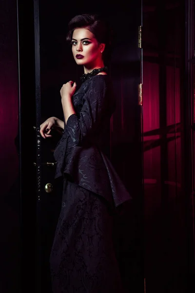 Portrait de beau mannequin de mode en robe noire classique, maquillage et coiffure près de porte sombre debout et posant et lumière rouge de la fenêtre brille sur elle . — Photo