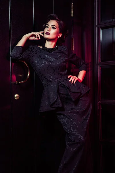 Retrato de modelo de moda bonita em vestido preto clássico, maquiagem e penteado perto da porta escura de pé e posando e luz vermelha da janela brilha sobre ela . — Fotografia de Stock
