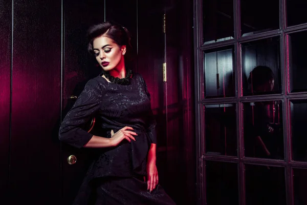 Portrait of beautiful fashion model in classic black dress, makeup and hairstyle near dark door standing and posing and red light from window glows on her. — Stock Photo, Image