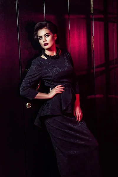 Retrato de modelo de moda hermosa en vestido negro clásico, maquillaje y peinado cerca de la puerta oscura de pie y posando y la luz roja de la ventana brilla en su . — Foto de Stock