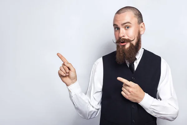 Bell'uomo d'affari con barba e baffi da manubrio che guarda la macchina fotografica, sorprende e mostra lo spazio di copia con le dita. studio girato, su sfondo grigio . — Foto Stock