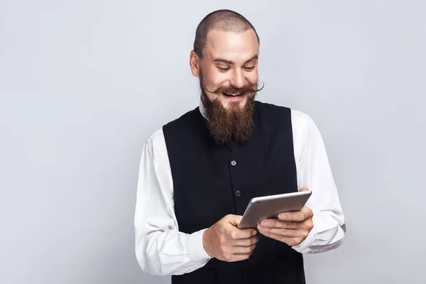 Stilig affärsman med skägg och mustasch håller digital tablett tittar på skärmen med toothy leende. Studio skott, på grå bakgrund. — Stockfoto